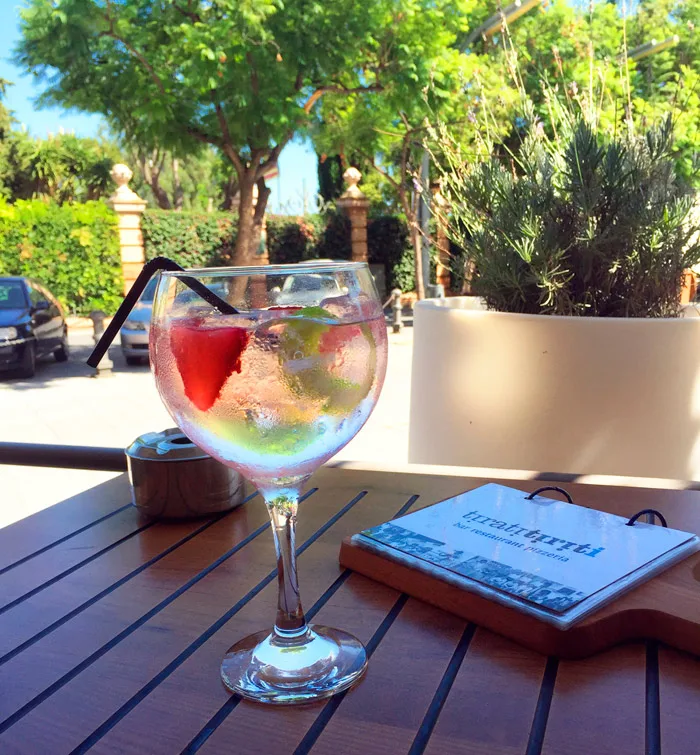 Copa de gin-tonic con fruta en la terraza, con los árboles de la plaza al fondo
